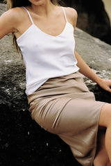 Close-up of a model wearing white silk camisole and taupe silk slip skirt posing on a rock by the beach