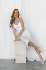 Model wearing white silk camisole and cream silk skirt posing on a marble plinth in a studio