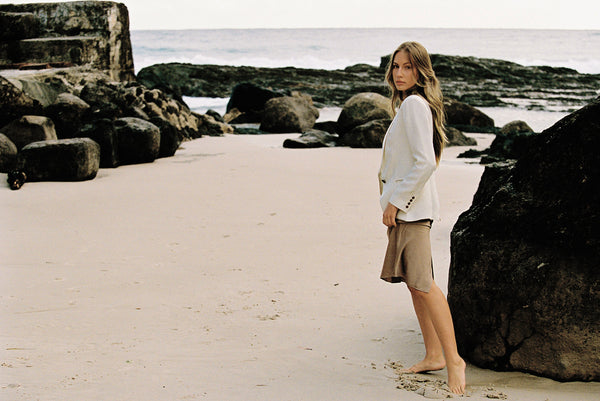 Woman wearing a white linen blazer and taupe silk skirt posing on a rock by the beach