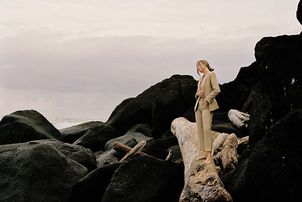 Woman wearing taupe linen blazer and pant sitting on a rock by the beach