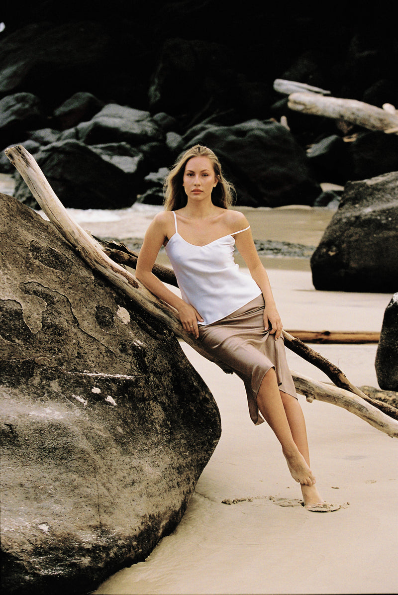 Woman wearing white silk camisole and taupe silk slip skirt leaning on a large rock at the beach