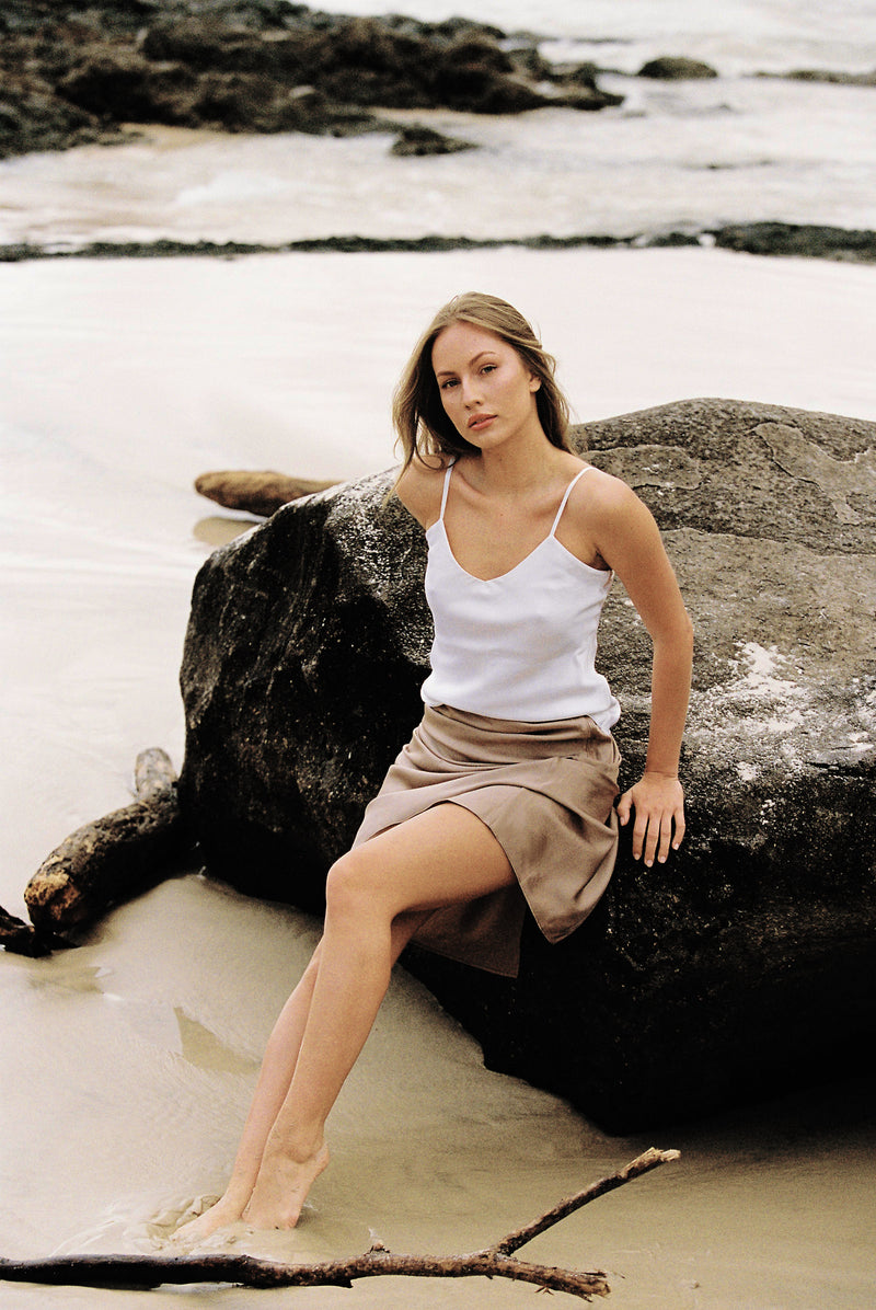 Woman wearing white silk camisole and taupe silk slip skirt sitting on a rock by the beach