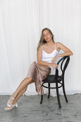 Model wearing a white silk camisole and taupe silk slip skirt posing on a chair in a studio