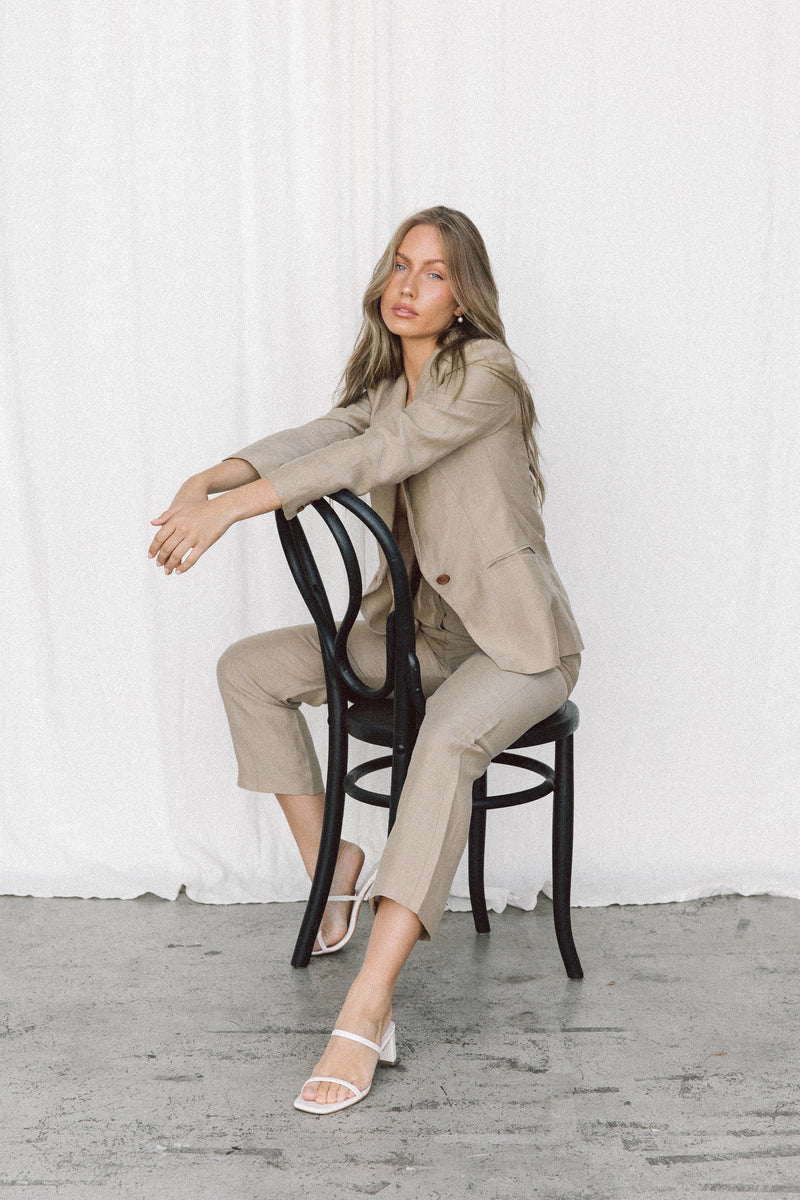 Model wearing a taupe blazer and trouser sitting backwards on a chair in a studio